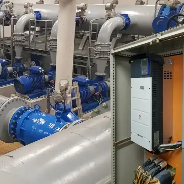 Großer Anlagenraum mit mehreren blauen Industriepumpen und Rohrleitungen; im Vordergrund ein offener Schaltschrank mit einem Frequenzumrichter.