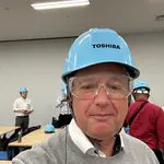 Thomas Dahmen von esco antriebstechnik trägt Sicherheitshelm und Schutzbrille während der Werksbesichtigung bei Toshiba in Tokio. Im Hintergrund bereiten sich weitere Teilnehmer auf den Rundgang vor.